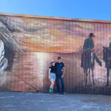 Angela stands with her partner in front of a shipping container with artwork of horses painted on it.