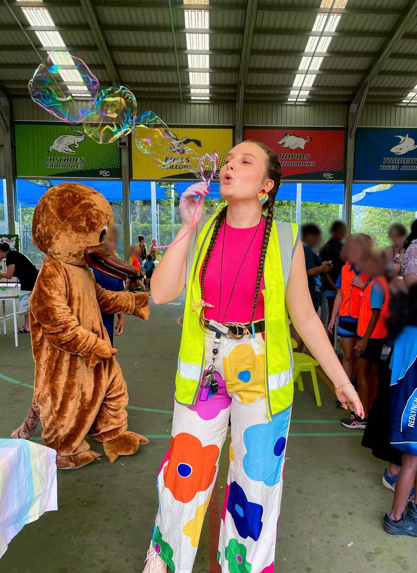 A teacher is standing in a school hall blowing bubbles. Her hair is in pig tails and she is wearing a bright pink top, a fluorescent yellow vest and white pants with big flowers on them.
