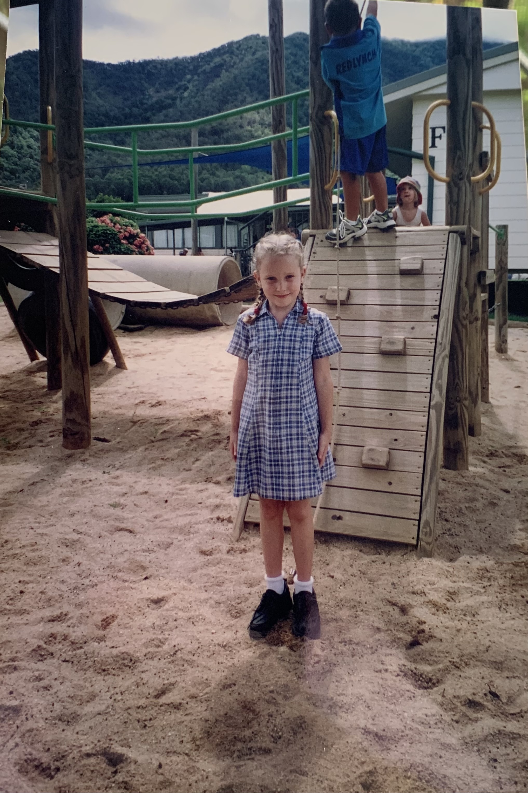 A child stands in a blue and white school uniform on a playground. She is smiling and her hair is in pigtails.
