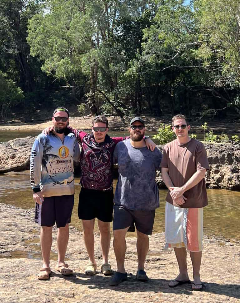 Photo of Declan and 3 friends hugging and smiling at the camera, in front of a stream and green trees.