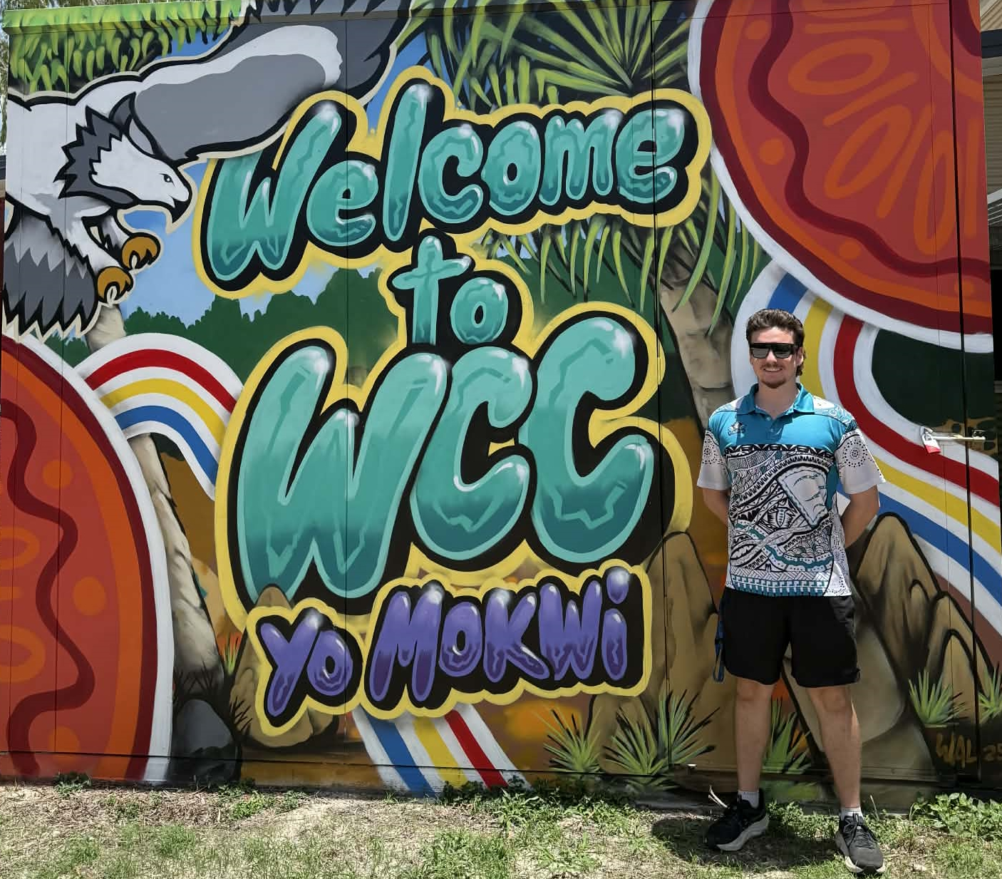 Declan standing smiling at the camera in front of a 'Welcome to WCC Yo MOKWi' mural with a bird of prey trees and bright colours