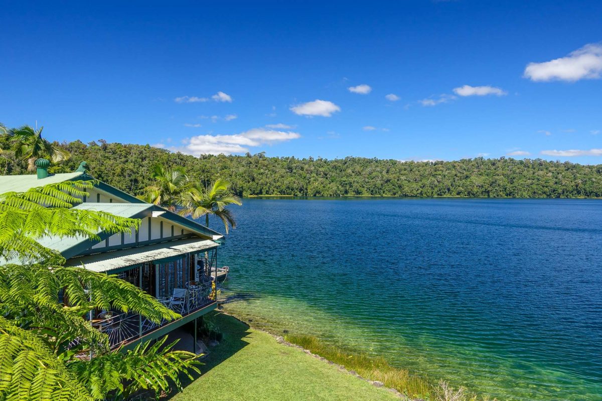 Photo of Cairns coast, with blue and green water and blue sky with little cloud. House on stilts and palm trees sit on the water