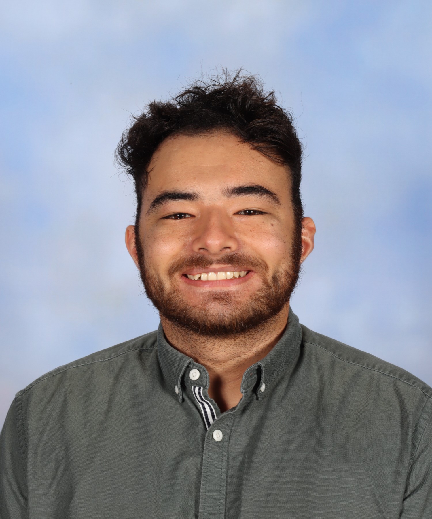 Photo of male teacher, Alex, with short dark curly hair and tan skin. He is smiling at the camera and wears a grey shirt.