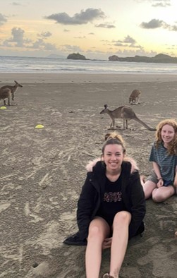 Photo of two women sitting on a beach, Cape Hillsborough, with 3 kangaroos and a sunset behind them.