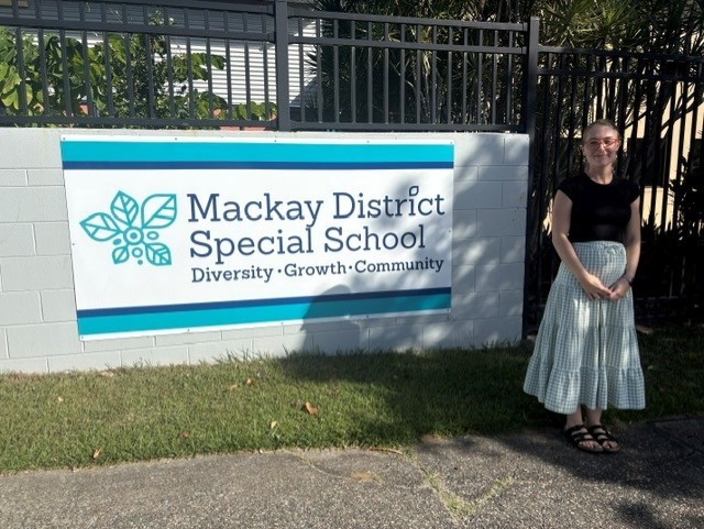 A photo of a teacher, Ashlee, standing beside a school sign that says "Mackay District Special School" in the sun.