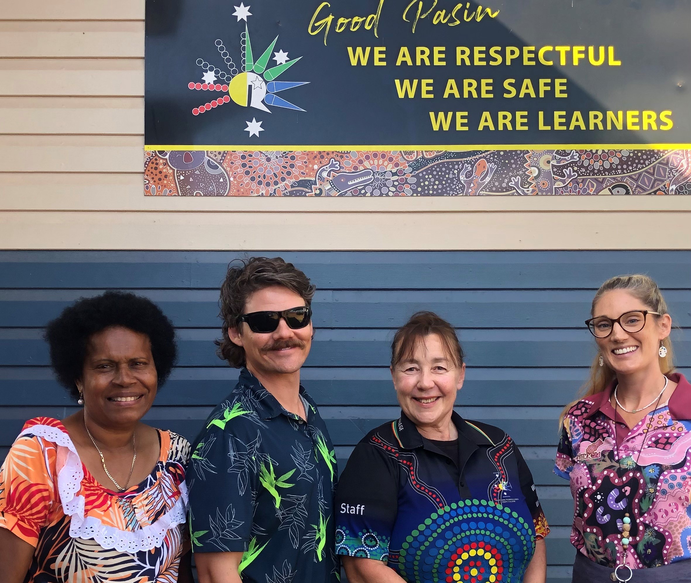 A group of four teachers smiling as they stand in front of a school sign that says, ‘We are respectful, we are safe, we are learners.’
