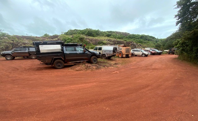 A group of off-road vehicles and campers are parked on a wide, red dirt lot, with a rocky hill in the background.